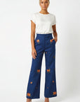 A Model in the Fanm Mon Cora wide-leg trousers with yellow and orange floral embroidery, styled with a white t-shirt and nude high-heeled sandals against a white studio background.