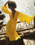 A woman wearing a bright yellow, knee-length, floral embroidery detail dress with a plunging neckline and bow tie front detail, standing by a body of water.