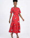 Model wearing the Liza Dress featuring jasmine-inspired floral embroidery, a buckle waist belt, short sleeves, and a gathered midi hem.
