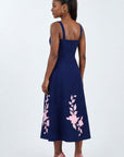 Model wearing the Fanm Mon Elmas Dress featuring oversized hibiscus-inspired embroidery, square back neckline, fitted bodice, and softly flared midi skirt against a neutral studio background.