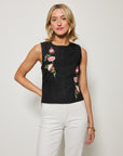 Woman wearing a black sleeveless top with floral embroidery and white pants on a plain background