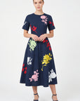 Model wearing Fanm Mon Gandi Dress in navy featuring a fitted waist, short sleeves, flowing A-line midi skirt, and pink, red, yellow, green, and white floral embroidery