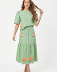 Model wearing Fanm Mon Komarna Set in green tea featuring a puff-sleeve top with scalloped edges, colorful floral embroidery, and a coordinating button-front midi skirt with patch pockets