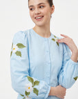 Close-up of Fanm Mon Paysan Dress in light blue showing white floral embroidery, green leaves, fabric-covered buttons, and gathered puff sleeves