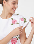 Close-up of Fanm Mon Pervin Dress in white highlighting the round neckline, short sleeve construction, and intricate pink rose embroidery with green leaves across the shoulder and bodice.