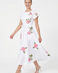 Model wearing Fanm Mon Pervin Dress in white with a round neckline, short sleeves, self-tie waist belt, and pink embroidered roses with green botanical foliage across the flowing midi skirt.