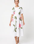 Model wearing Fanm Mon Rumen Dress in white with a round neckline, short sleeves, defined waist, and oversized pink rose embroidery with green foliage across the flowing midi skirt.