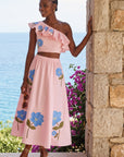 Model wearing the Fanm Mon Tamira Set in light pink featuring a one-shoulder ruffled crop top and flowing midi skirt with blue floral embroidery