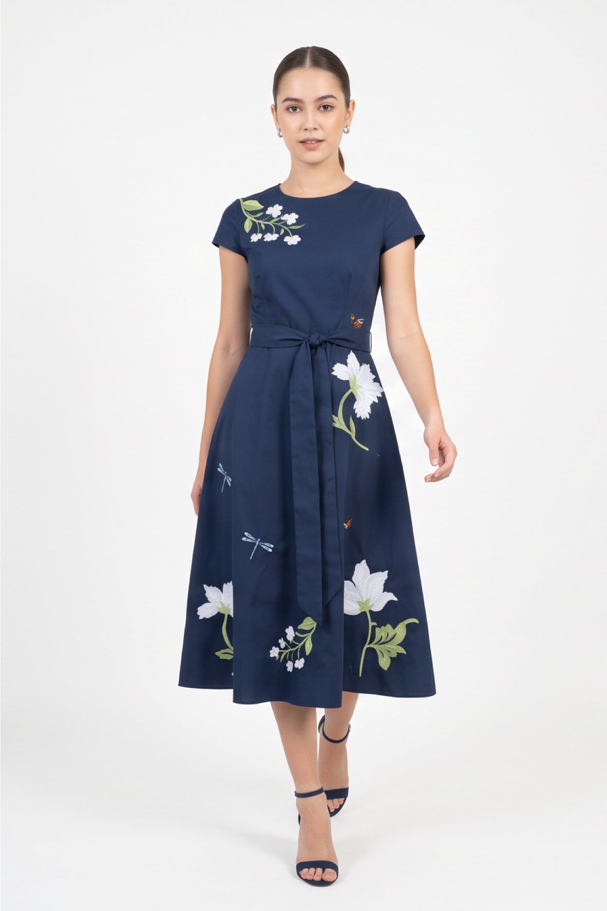Model wearing the Fanm Mon Burma Dress featuring a crew neckline, short sleeves, removable self-tie waist belt, embroidered anemones, jasmine blossoms, dragonflies, butterflies, and an A-line midi skirt.