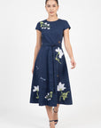 Model wearing the Fanm Mon Burma Dress featuring a crew neckline, short sleeves, removable self-tie waist belt, embroidered anemones, jasmine blossoms, dragonflies, butterflies, and an A-line midi skirt.