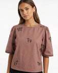 Model wearing Fanm Mon Cait short-sleeve blouse with black embroidered horse motifs, paired with black pants, against a plain white background.
