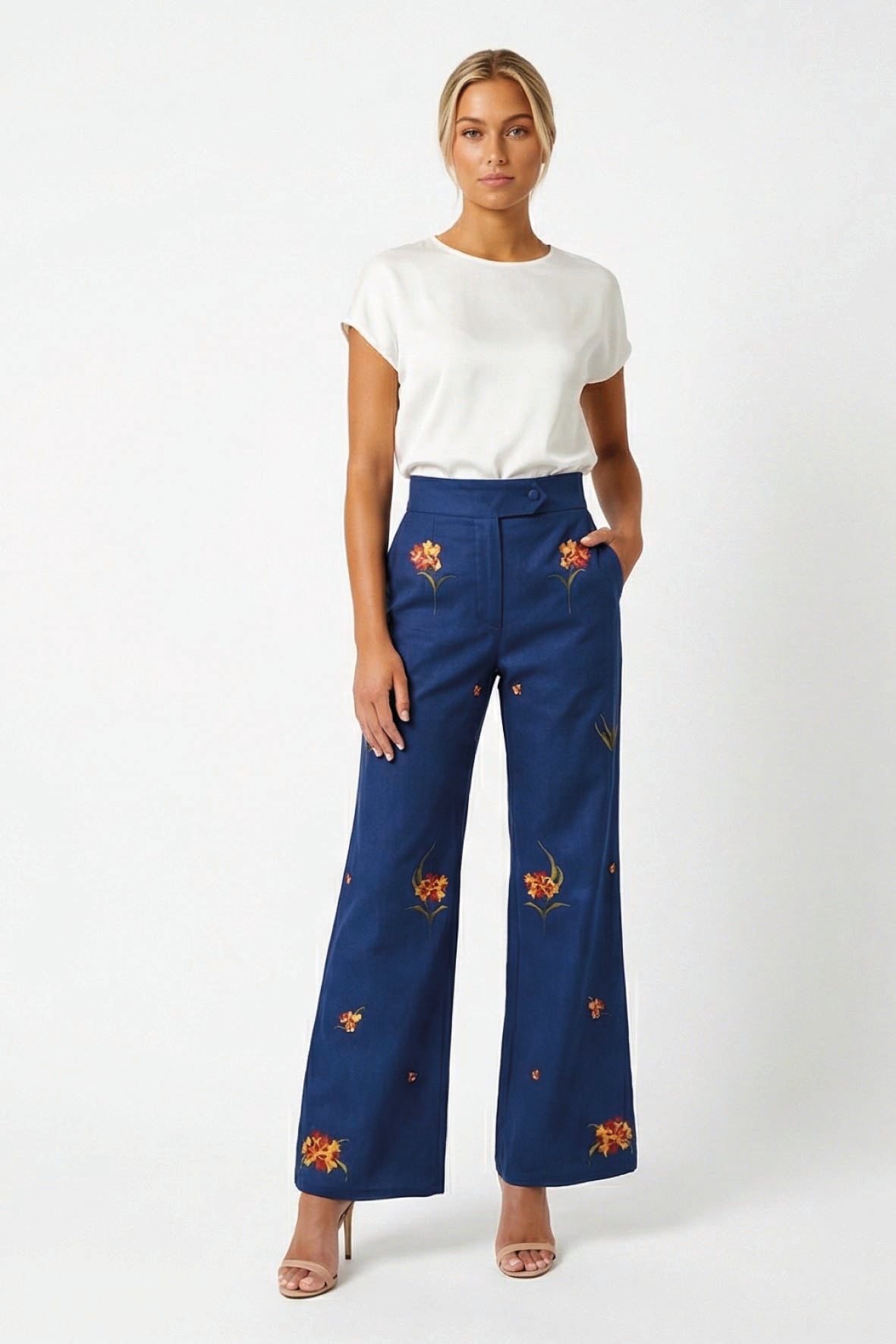 A Model in the Fanm Mon Cora wide-leg trousers with yellow and orange floral embroidery, styled with a white t-shirt and nude high-heeled sandals against a white studio background.