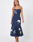 Model wearing the Fanm Mon Gayelle Dress featuring cosmos flower embroidery, cherry blossom details, dragonflies, and bee motifs with a fitted bodice and flounced midi hem against a neutral background.