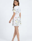 Model wearing the Fanm Mon Joe Dress in White with trailing wildflower embroidery, short sleeves, and tailored mini silhouette shown from the back against an off white background.