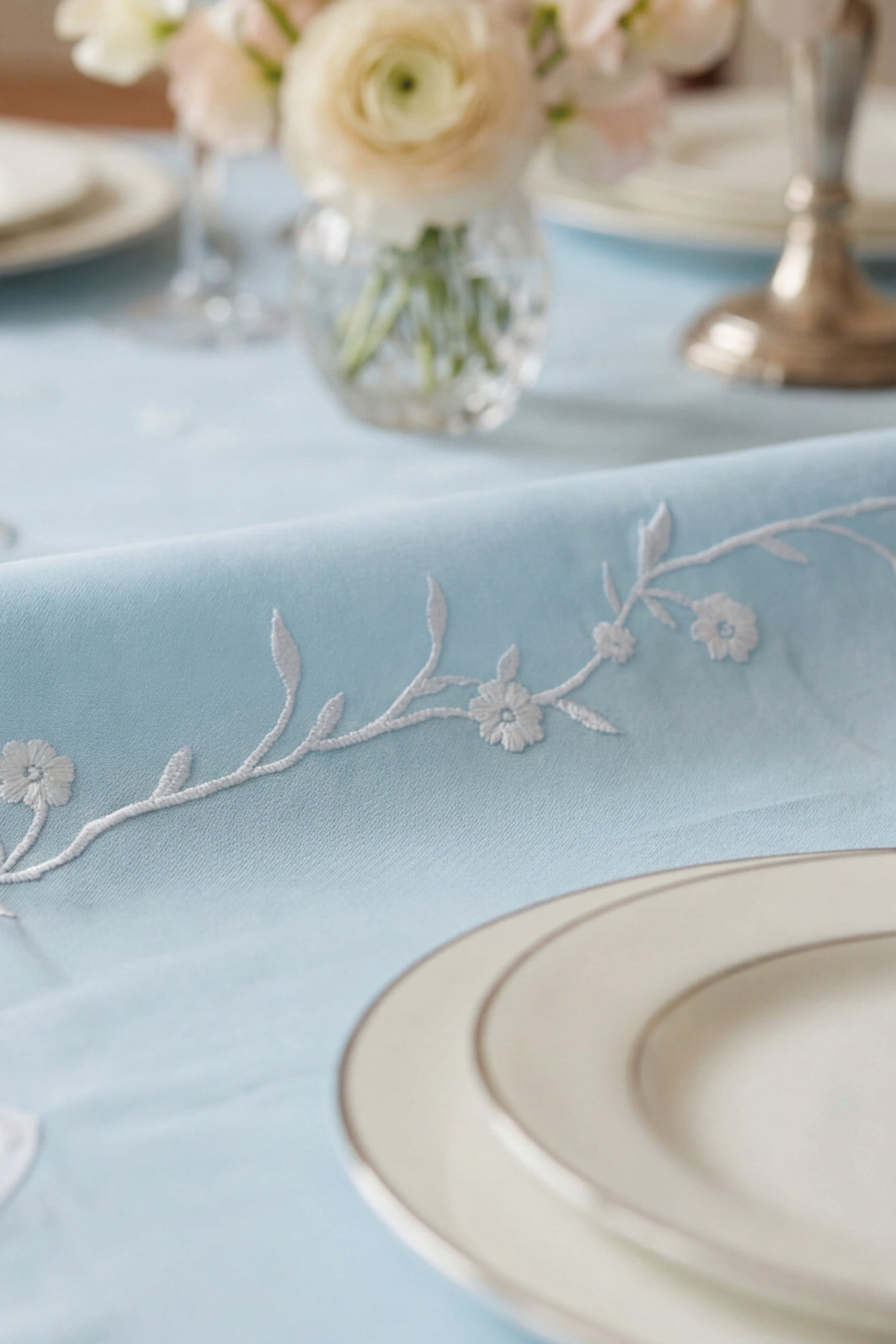Light blue tablecloth with floral embroidery and a white plate on a table setting.