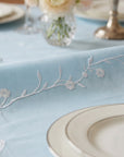 Light blue tablecloth with floral embroidery and a white plate on a table setting.