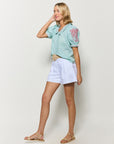Model wearing Fanm Mon’s Puren Top with a scalloped collar and button-down front, in green check plaid with pink floral embroidery on the sleeves and collar. The top is styled with white shorts against a plain background.