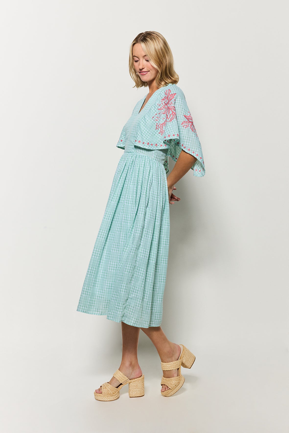 Side view of the Model wearing Fanm Mon's Tunus Midi Dress in Green Check plaid with pink floral embroidery on a white background