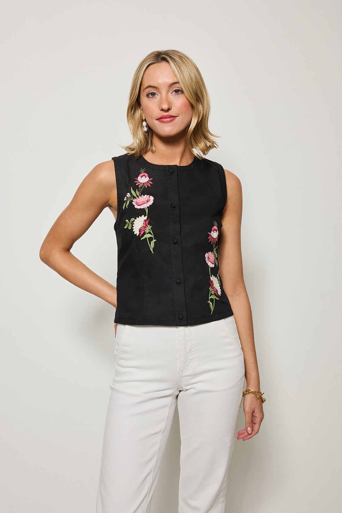 Woman wearing a black sleeveless top with floral embroidery and white pants on a plain background