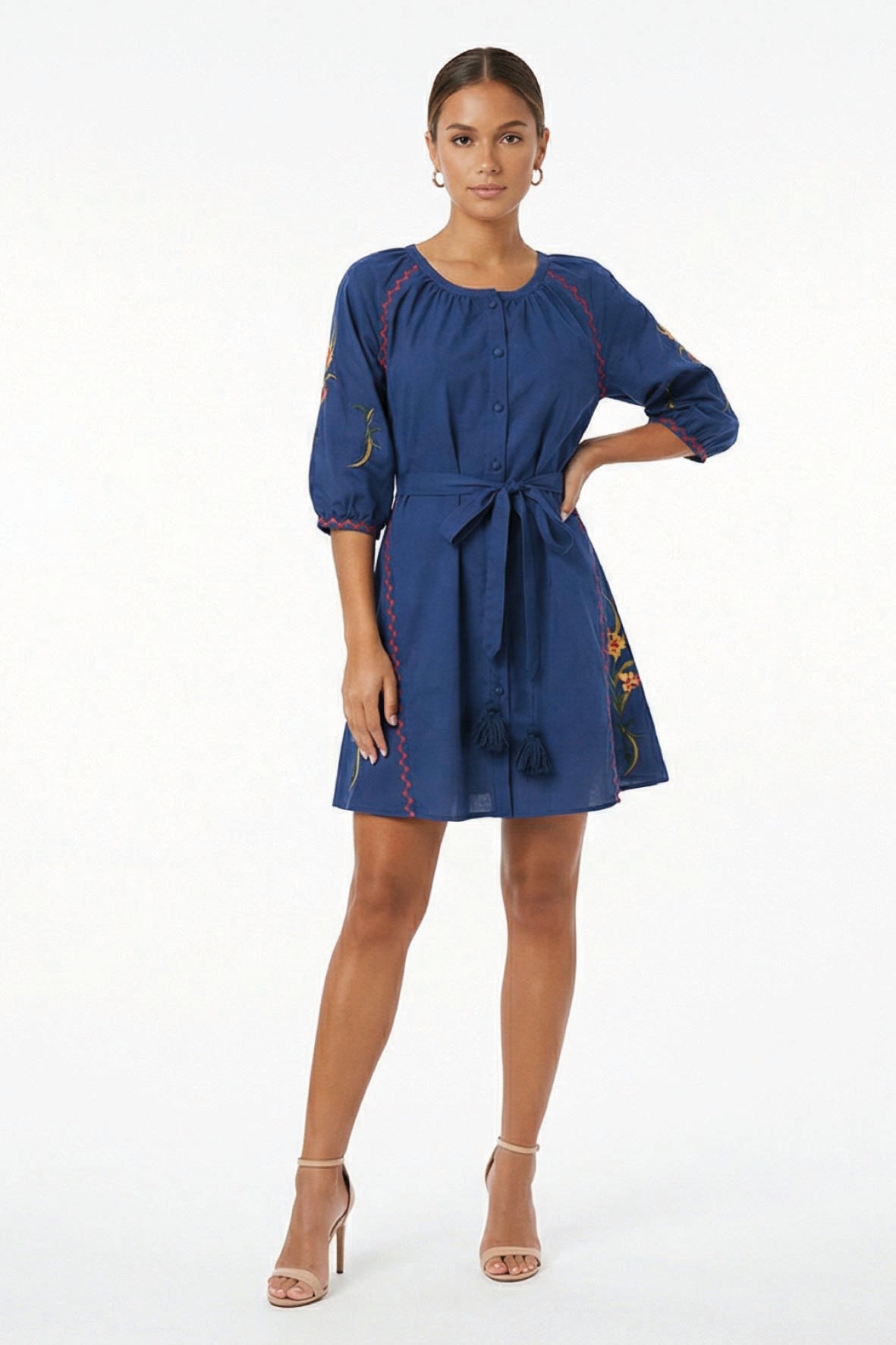 Model wearing the Fanm Mon Pearl Dress featuring a gathered round neckline, fabric-covered button front, three-quarter balloon sleeves, self-tie waist, tassel details, and floral embroidery.