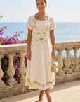 Model wearing the Fanm Mon Selma Dress featuring a square neckline, short sleeves, decorative covered buttons, embroidered floral motifs, peplum waist, and tiered midi skirt overlooking the coast.