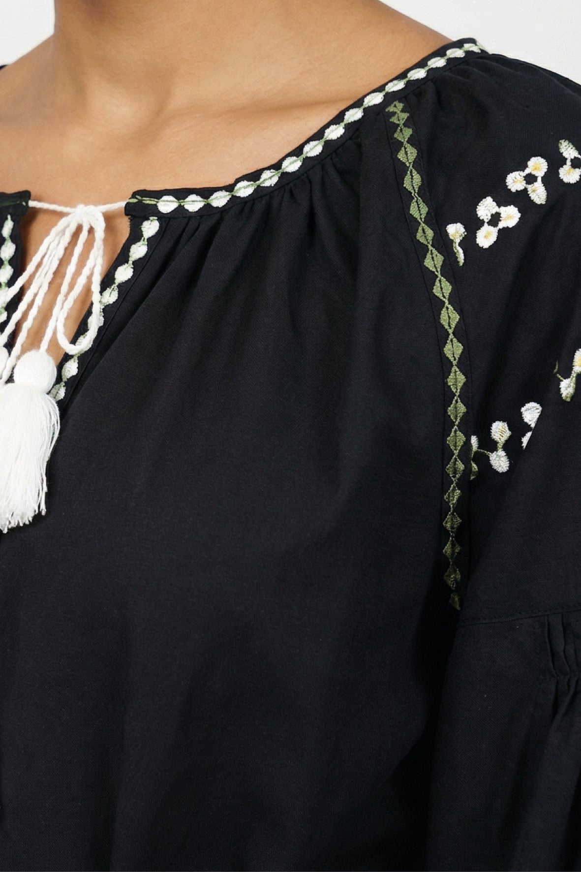 Close-up of a black Stella Dress with white tassel tie, white floral embroidery, and green geometric trim on the neckline and sleeve.