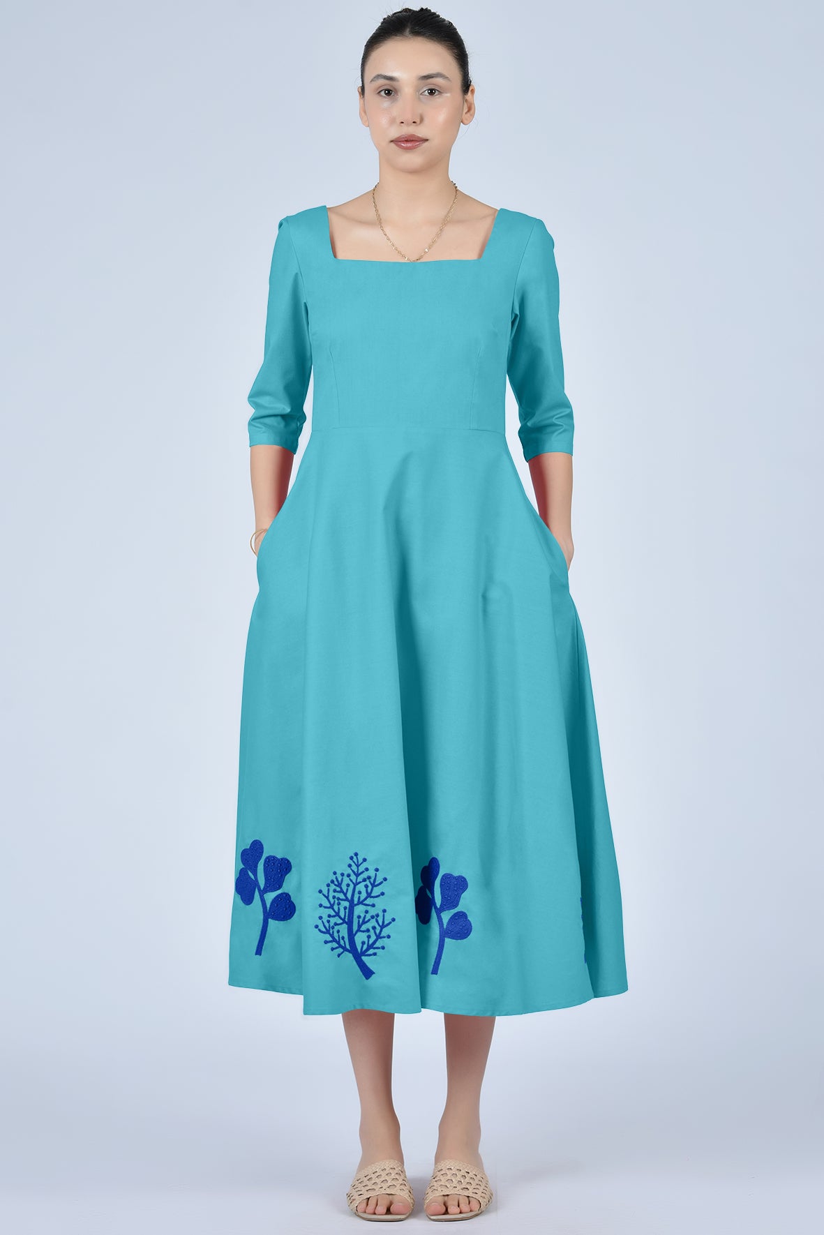 Woman wearing a teal dress with floral embroidery on a light gray background