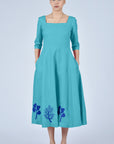 Woman wearing a teal dress with floral embroidery on a light gray background
