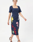 Model wearing the Fanm Mon Blanka Dress in indigo blue with a tailored bodice, square neckline, and embroidered yellow, pink, red, white, and green botanical motifs.