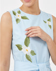 Close-up of Fanm Mon Gemimi Dress in light blue highlighting the round neckline, sleeveless bodice, white embroidered flowers with yellow centers, green foliage, and coordinating waist tie.