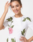 Close-up of Fanm Mon Rumen Dress in white highlighting the round neckline, short sleeves, pink rose embroidery with red detailing, and intricate green botanical foliage across the fitted bodice.