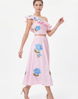 Model wearing the Fanm Mon Tamira Set in light pink with a ruffled one-shoulder crop top, gathered A-line midi skirt, and oversized blue floral embroidery